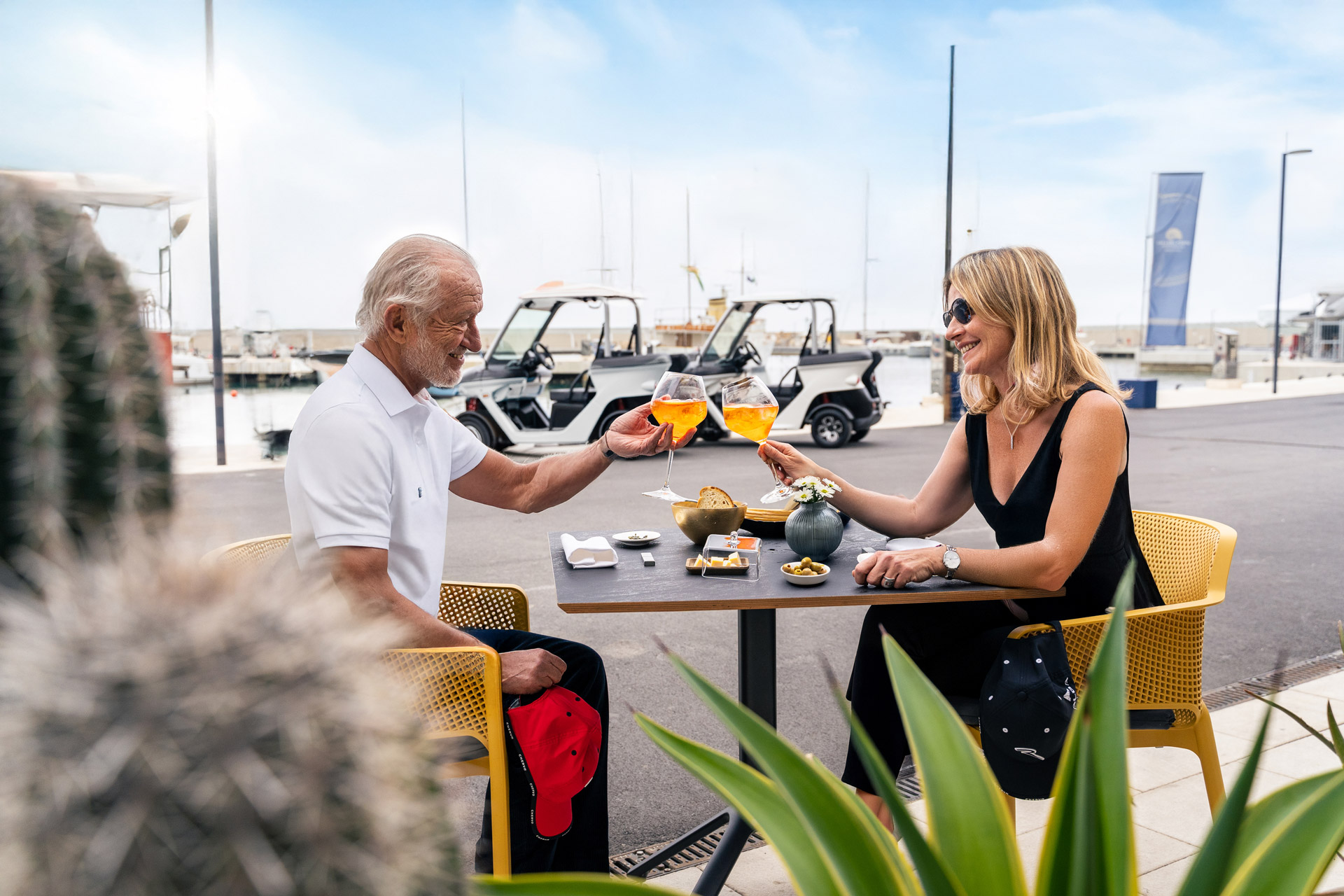 In primo piano una coppia che si presta a consumare un aperitivo dove aver parcheggiato due Alba 2SC sullo sfondo lungo la banchina del porto.