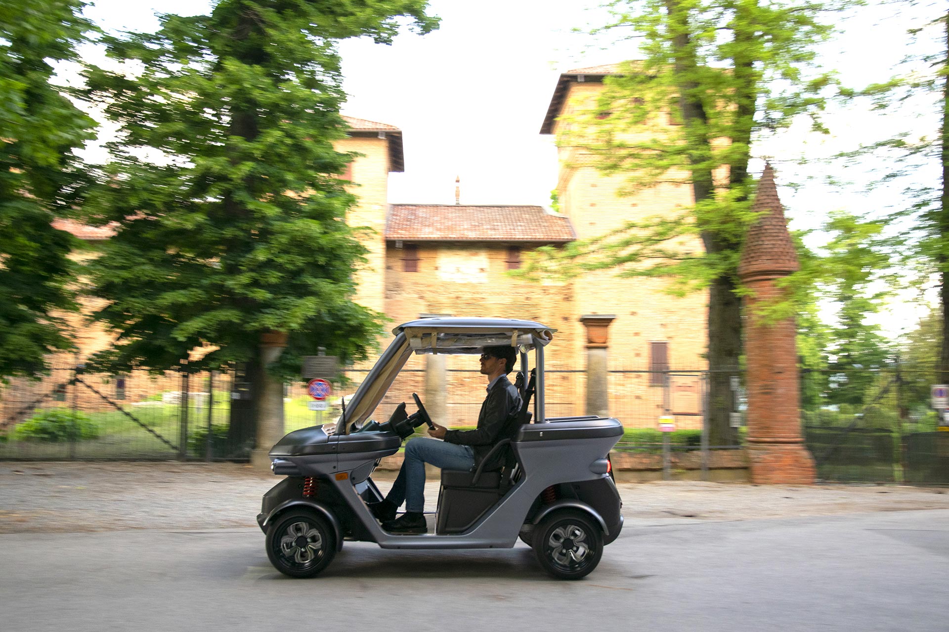 Alba 2SC in movimento su una strada urbana, guidato da una persona, con un edificio storico in mattoni e alberi sullo sfondo.