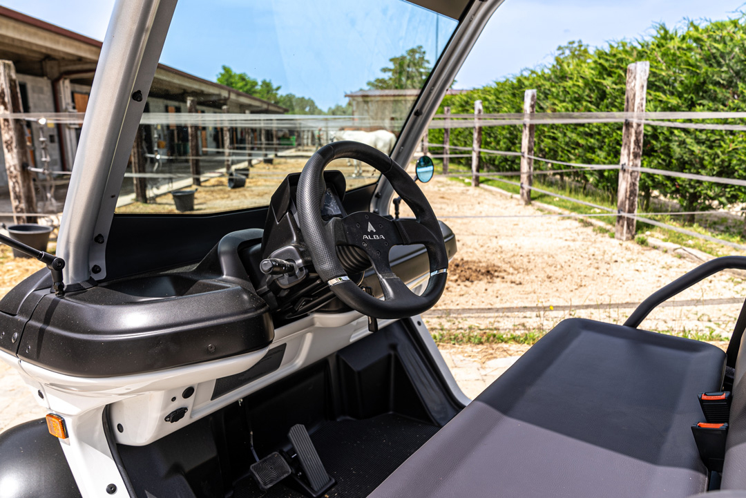 Interno di un Alba 2SC CARGO con volante e sedile in primo piano, parcheggiato in un’area rurale con recinti e strutture di un maneggio sullo sfondo.