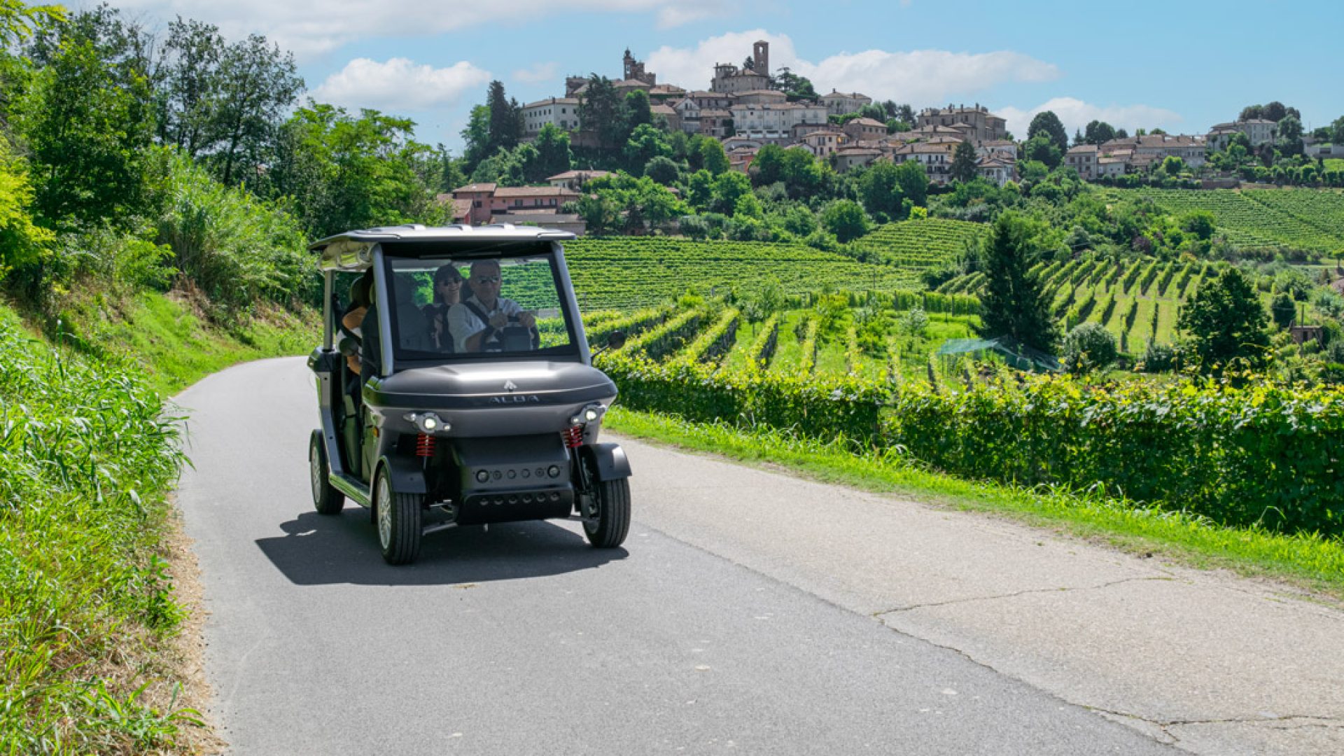Cart elettrico 4 posti stradale Alba 4SC, con guida NCC e taxi, che percorre una strada collinare turistica tra vigneti verdi, con un borgo sullo sfondo.